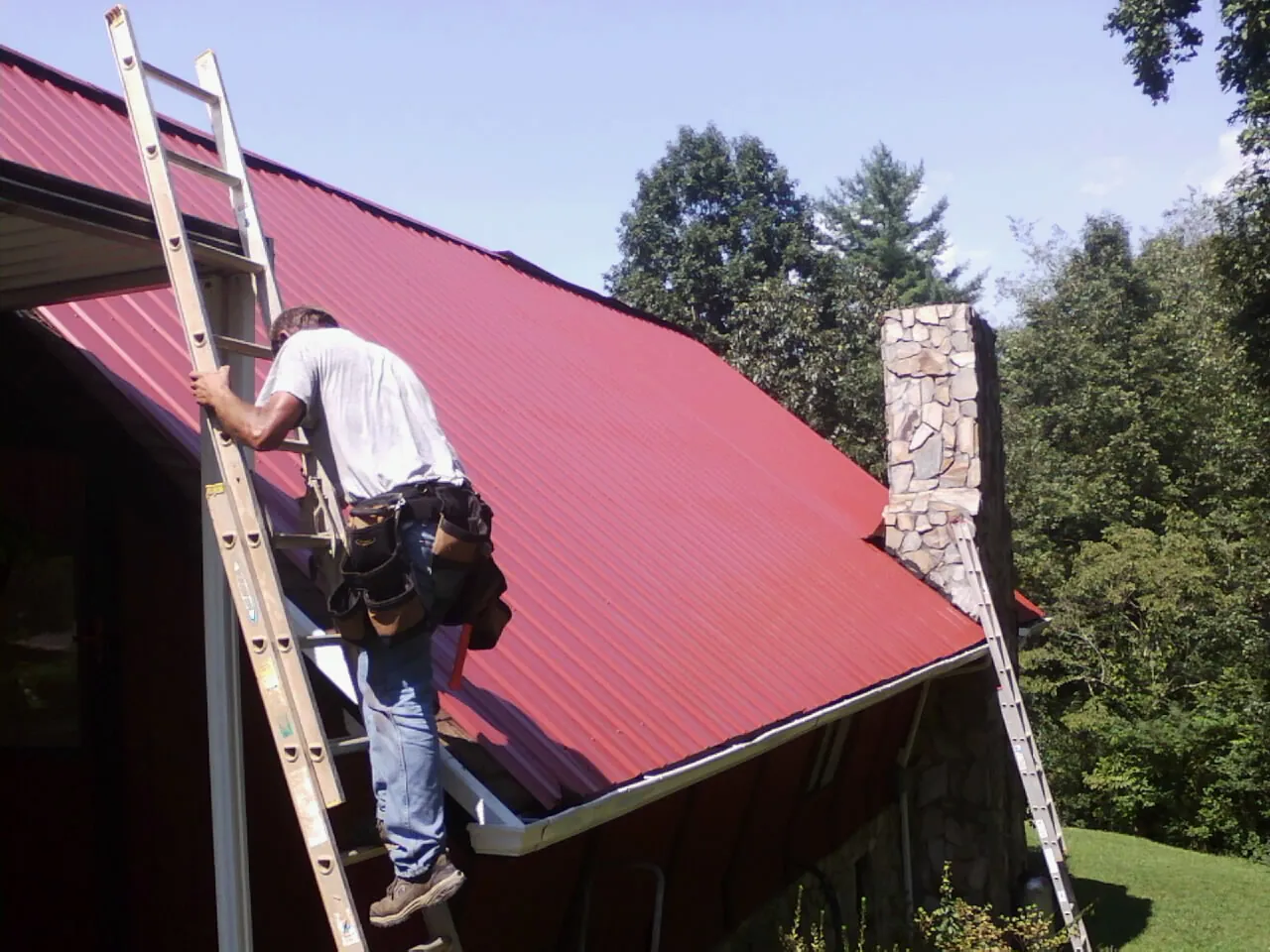 Roofer on a ladder beside a red metal roof and stone chimney — Soffit & Fascia Repair by Roofing Brothers in Neosho